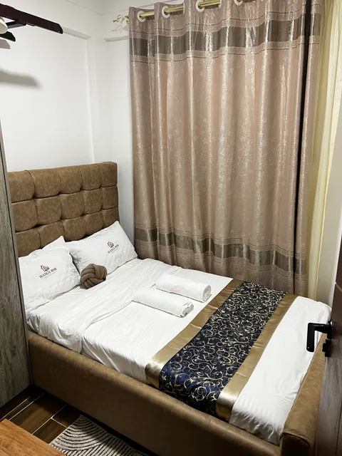 Luna bedroom with an upholstered headboard, white bedding, rolled towels and patterned curtains.