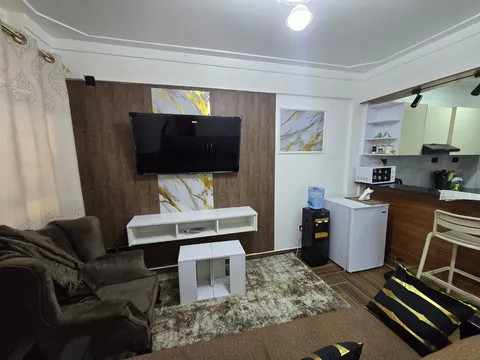 Sol living room with a wall-mounted television, white coffee tables, armchair and brown sofa beside the open kitchen and breakfast counter.